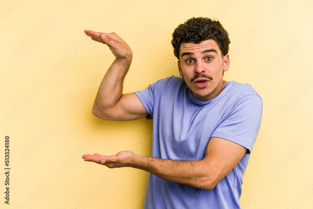 Young caucasian man isolated on yellow background shocked and amazed holding a copy space between hands. Young caucasian man isolated on yellow background shocked and amazed holding a copy space between hands.