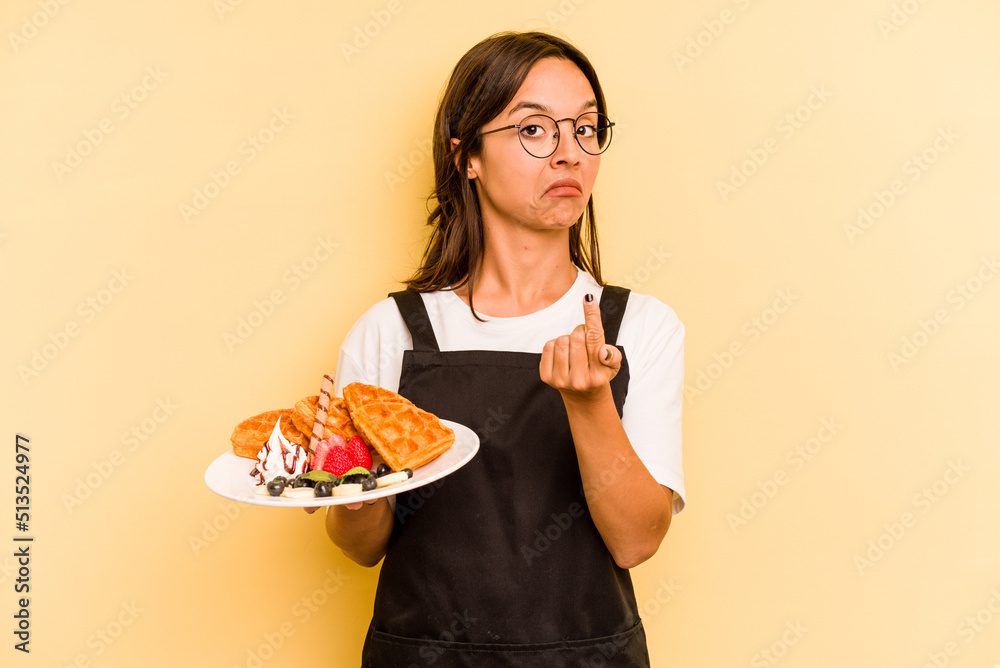 Young hispanic dependent woman holding waffles isolated on yellow background pointing with finger at you as if inviting come closer. Young hispanic dependent woman holding waffles isolated on yellow background pointing with finger at you as if inviting come closer.