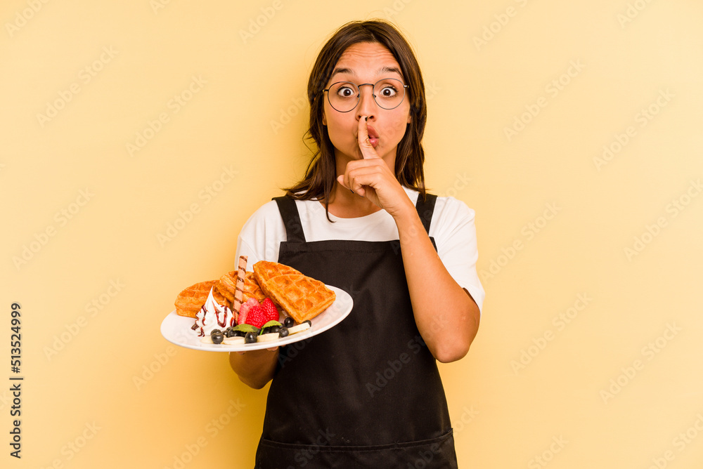 Young hispanic dependent woman holding waffles isolated on yellow background keeping a secret or asking for silence.