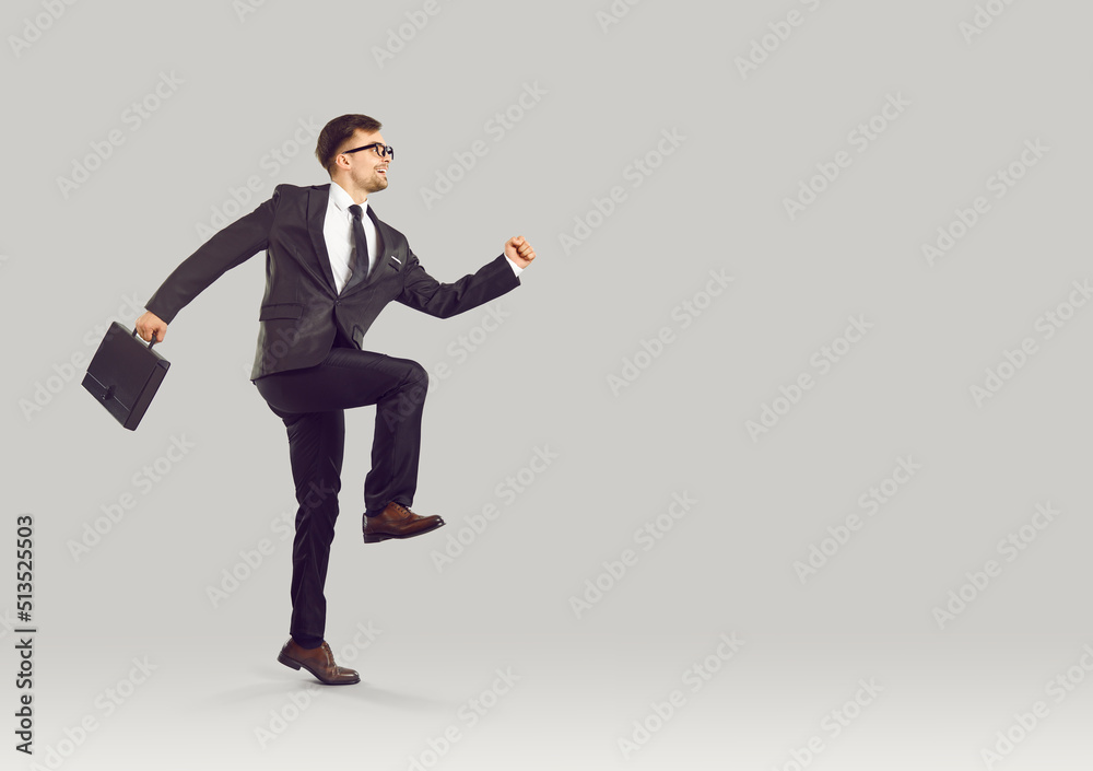 Strange happy office worker holding business briefcase and running to work in the morning. Full body length side profile view of funny man in suit running and skipping on light gray studio background