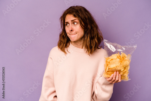 Young caucasian woman holding a bag of chips isolated on purple background confused  feels doubtful and unsure.