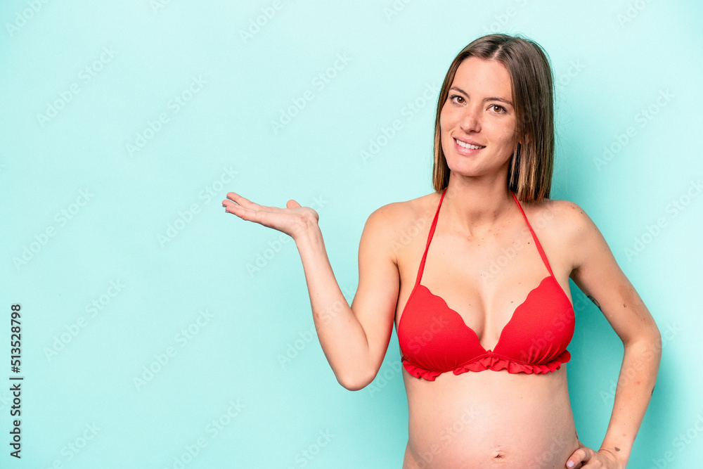 Young caucasian pregnant woman wearing bikini isolated on blue background showing a copy space on a palm and holding another hand on waist. Young caucasian pregnant woman wearing bikini isolated on blue background showing a copy space on a palm and holding another hand on waist.