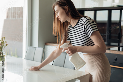 Portrait of beautiful young woman cleaning white table with spray and cloth on modern kitchen side view. Housekeeper tidy up kitchen furniture with cleanser. Clean and beautiful home