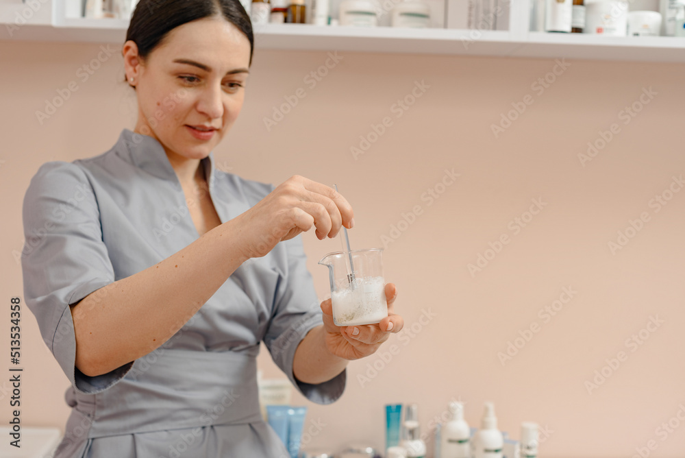 Young woman dermatologist in gray uniform mixing white cream in glass jar Young woman dermatologist in gray uniform mixing white cream in glass jar