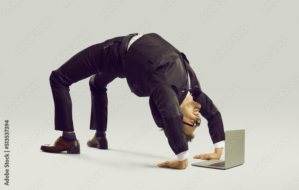 Happy energetic businessman practises yoga positions while working on  laptop computer. Funny fit flexible young man in suit and glasses doing  bridge pose on studio floor while using modern notebook PC Photos