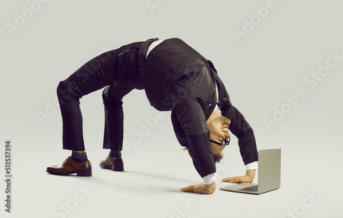 Happy energetic businessman practises yoga positions while working on laptop computer. Funny fit flexible young man in suit and glasses doing bridge pose on studio floor while using modern notebook PC photo