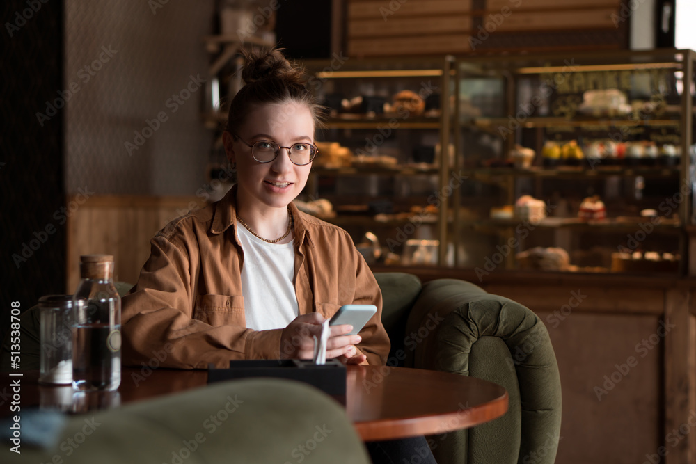 Young smiling woman sitting in cafe drinking coffe and using smartphone