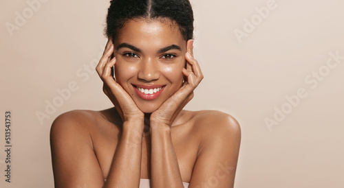 Smiling african american woman, applies daily care, nourishing facial mask on her skin and looking happy, standing over brown background