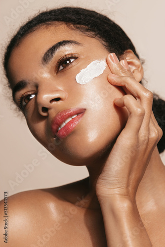 Vertical of beauty woman, dark skinned model applies facial cream, skincare mask treatment for face, stands over brown background photo