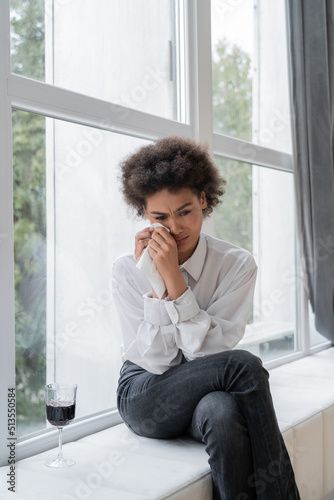 Wallpaper Mural sad african american woman wiping tears near glass of red wine while sitting on windowsill. Torontodigital.ca