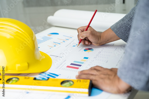 Asian engineer with architectural house plan project blueprint, graph and yellow helmet and engineering construction tools.
