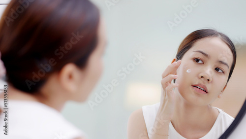 Fresh healthy skin of Asian girl looking at mirror, applying facial moisturizer on her face, putting cream treatment before makeup cosmetic.