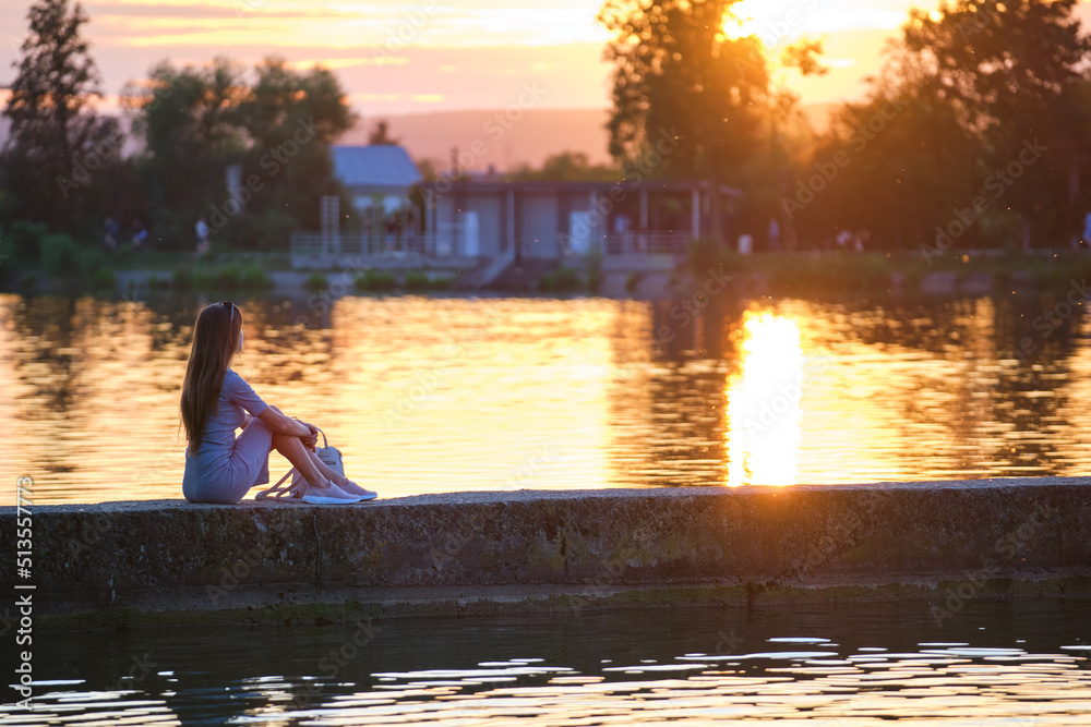 Back view of lonely young woman sitting alone on lake shore enjoying warm evening. Wellbeing and relaxing in nature concept Back view of lonely young woman sitting alone on lake shore enjoying warm evening. Wellbeing and relaxing in nature concept