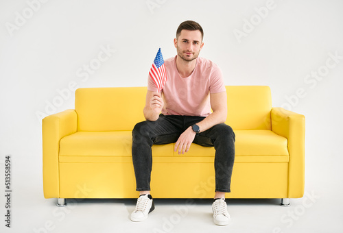 Young cheerful man with american flag in his hand sitting on bright sofa cover white background