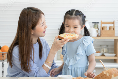 Happy Asian family  cute little daughter eating bread with jam  rainbow sugar flakes feeding from young mother after spreading jam on sliced bread together  preparing breakfast in kitchen at home