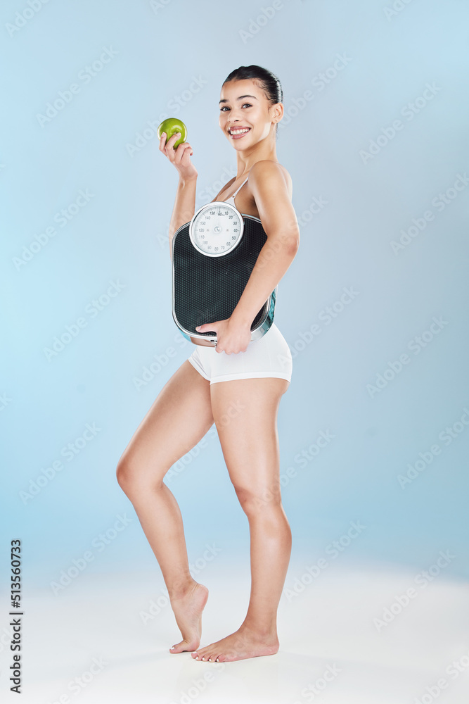 Beautiful woman holding scale and apple against blue studio background with copyspace. Portrait of one fit and happy young female showing weightloss progress from healthy lifestyle and balanced diet Beautiful woman holding scale and apple against blue studio background with copyspace. Portrait of one fit and happy young female showing weightloss progress from healthy lifestyle and balanced diet