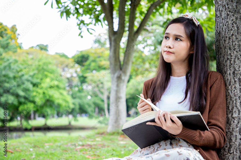 Smiling of Asian woman thinking after reading lessons and writing research in park. Reading the book in a garden during beautiful day. Smiling of Asian woman thinking after reading lessons and writing research in park. Reading the book in a garden during beautiful day.