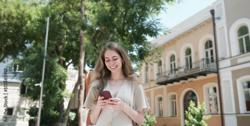 Young woman using smartphone outdoors. Joyful smiling woman girl looking at mobile phone in a city. Communication, technology, connection, lifestyle, summer vacation and travel concept