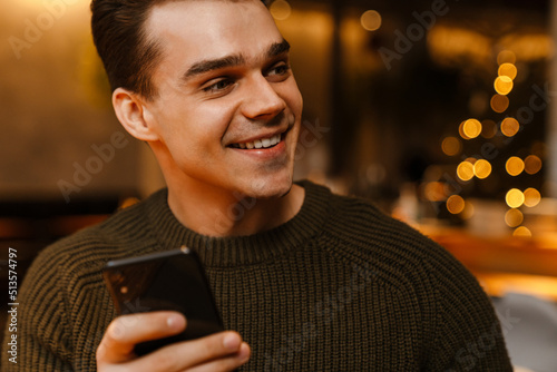 Young white man wearing sweater smiling while using cellphone in cafe