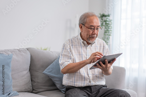 Asian grandfather wearing eyeglasses using digital tablet sitting on couch in the living room. senior man learning to use gadget technology and connecting online with family. 