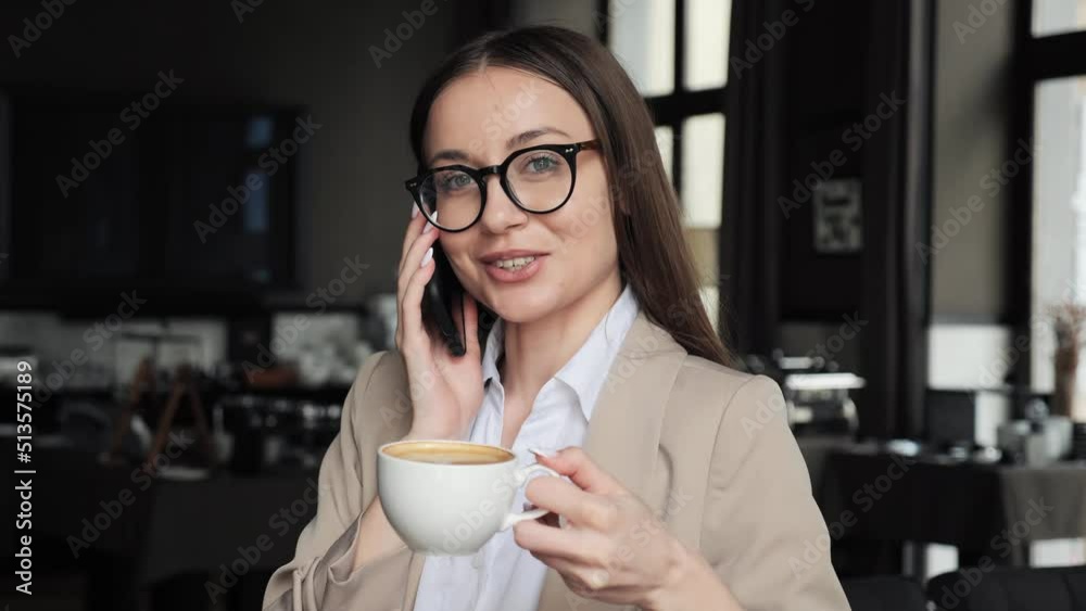 Stylish business woman in eyeglasses having mobile phone call while drinking coffee at hotel lounge restaurant. Businesswoman drinking cappuccino cup at cafe. Beautiful lady relaxing at office lobby. Stylish business woman in eyeglasses having mobile phone call while drinking coffee at hotel lounge restaurant. Businesswoman drinking cappuccino cup at cafe. Beautiful lady relaxing at office lobby.