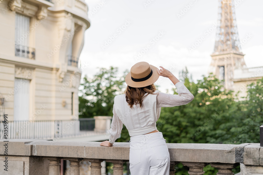 Back view of stylish woman holding sun hat with Eiffel tower at background in Paris.