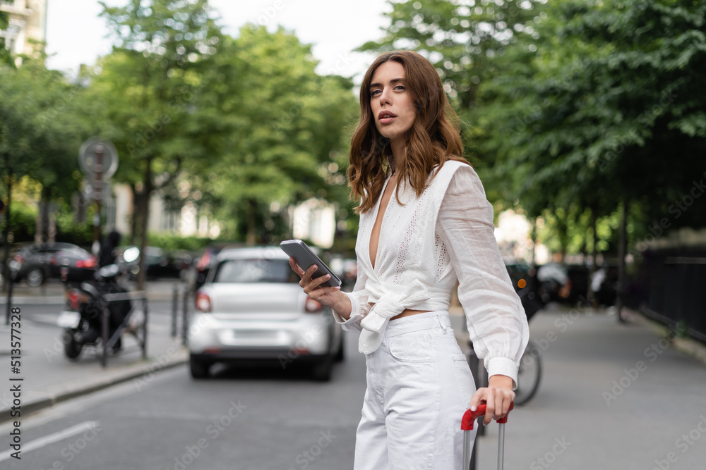 Stylish woman with cellphone and suitcase standing near road on street in Paris.