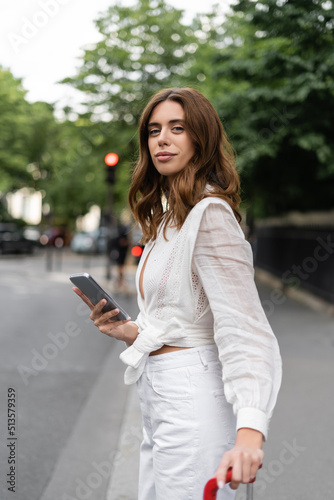Stylish brunette woman holding smartphone and suitcase on street in Paris.