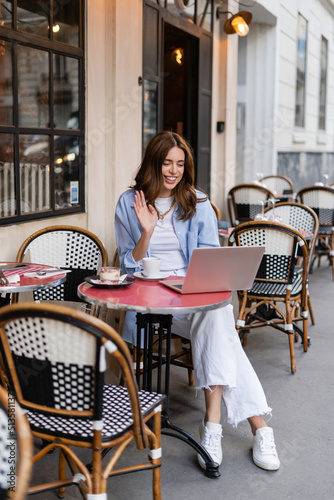 Smiling woman having video call on laptop near coffee in outdoor cafe in France. © LIGHTFIELD STUDIOS