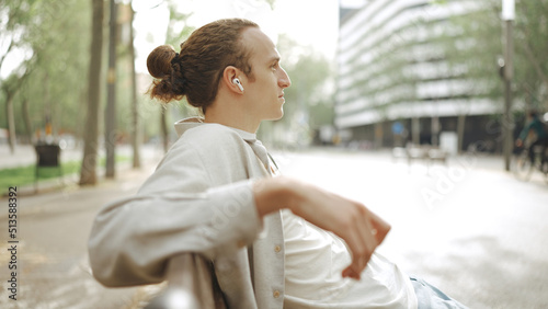 Young white man using earphones while sitting on bench outdoors