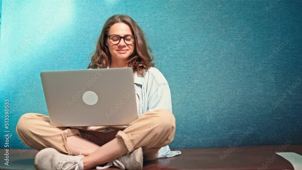 A young cheerful woman in glasses is typing on a laptop while sitting cross-legged on a couch on campus.