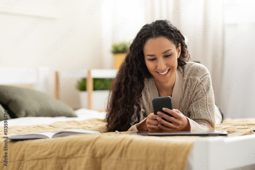 Happy young woman using mobile phone, having online call, lying on bed, posting in social media at home, copy space Happy young woman using mobile phone, having online call, lying on bed, posting in social media at home, copy space