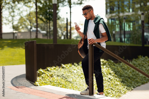 Stylish african american man in sunglasses holding smartphone while walking in urban area, texting or using navigator