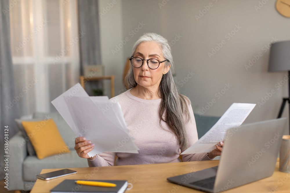 Serious european old female with gray hair in glasses works on computer and documents in room interior Serious european old female with gray hair in glasses works on computer and documents in room interior