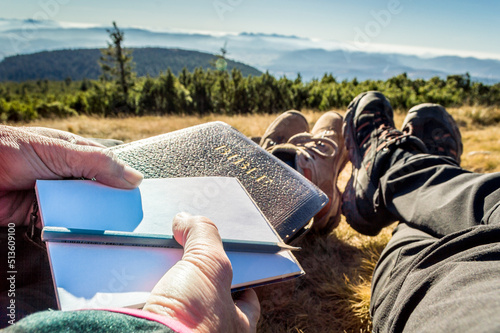 Outdoor Bible study during mountain hike in the fall. A couple sitting on the ground with legs stretched, holding the Bible, notepad and pencil. Mountaintop outlook on a hilly landscape.