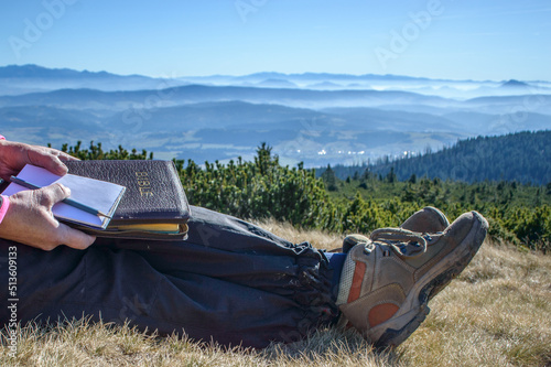 Outdoor Bible study during mountain hike. Female hands holding a Bible on the knees during outdoor devotion and Bible study in the mountains. Mountaintop outlook with high sky. Copy space.