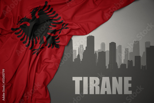 abstract silhouette of the city with text Tirane near waving national flag of albania on a gray background.