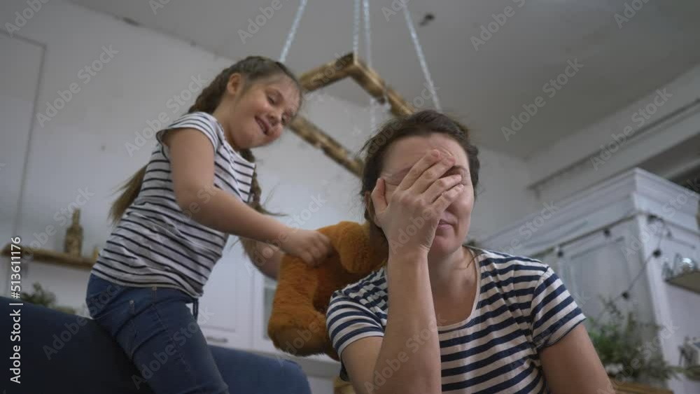 Irritated girl with sore head. Daughter screams and plays with teddy bear. Loud screams annoy sick mother in quarantine. Screaming of children disturbs parent. Mom with sore head is in quarantine. Irritated girl with sore head. Daughter screams and plays with teddy bear. Loud screams annoy sick mother in quarantine. Screaming of children disturbs parent. Mom with sore head is in quarantine.