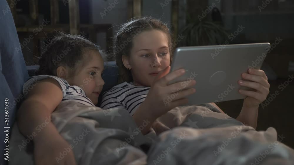 Children watch tablet lying on couch under warm blanket. Children watch movie on digital tablet secretly from adults while they sleep. children secretly play tablet under covers on couc Children watch tablet lying on couch under warm blanket. Children watch movie on digital tablet secretly from adults while they sleep. children secretly play tablet under covers on couc