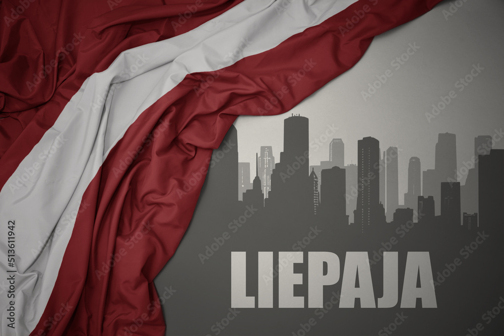 abstract silhouette of the city with text Liepaja near waving national flag of latvia on a gray background.