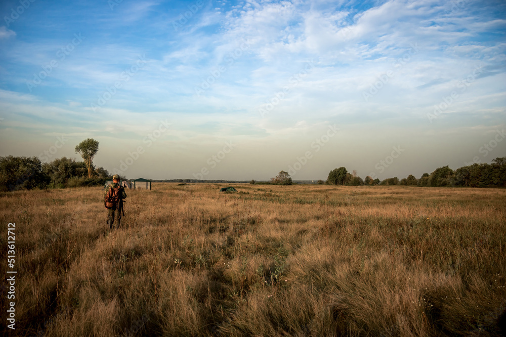 Hunting scene with hunter man in camouflage with shotgun going through rural field towards hunting camp during hunting season 