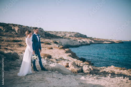 A young husband in a suit and a beautiful, slender bride in a wedding dress on a rocky seashore at sunset.