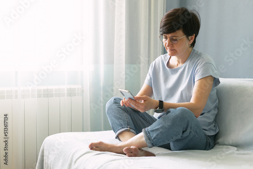 Middle-aged woman in glasses at home on the couch holding a mobile phone, online communication