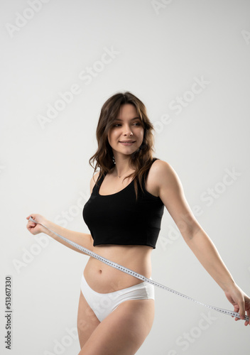 Cheerful beautiful sexy brunette woman girl in black top and white shorts with a measuring tape on a white background.