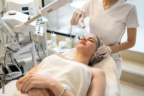 Beautician applying electric Darsonval facial massage treatment with special tool over female patients forehead in beauty salon photo