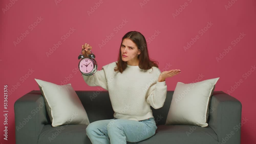 Hot deadline. Strict young woman holding alarm clock pointing by finger on time of clock face, sitting on sofa. Hurry up Hot deadline. Strict young woman holding alarm clock pointing by finger on time of clock face, sitting on sofa. Hurry up