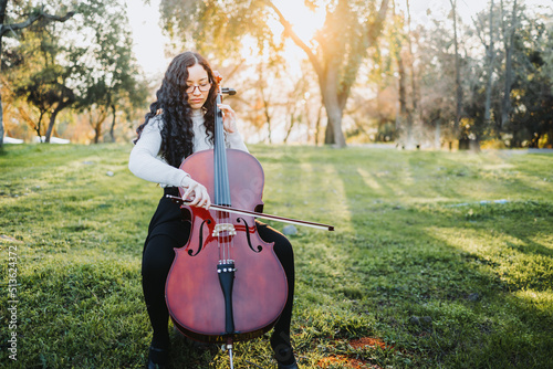 Young brunette woman with glasses playing cello at sunset in the park, on a green grass. Copy space.