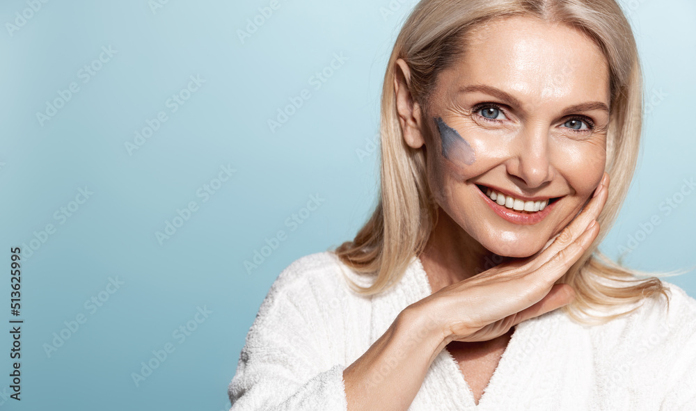 Naklejka premium Rejuvenation and skincare. Beautiful middle aged woman, 50 years, apply scrub, facial cream, smiling at camera, standing over blue background