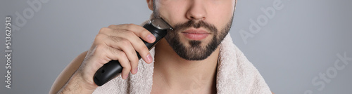 Handsome young man shaving with electric trimmer on grey background, closeup. Banner design photo