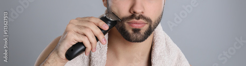 Fotografi Handsome young man shaving with electric trimmer on grey background, closeup